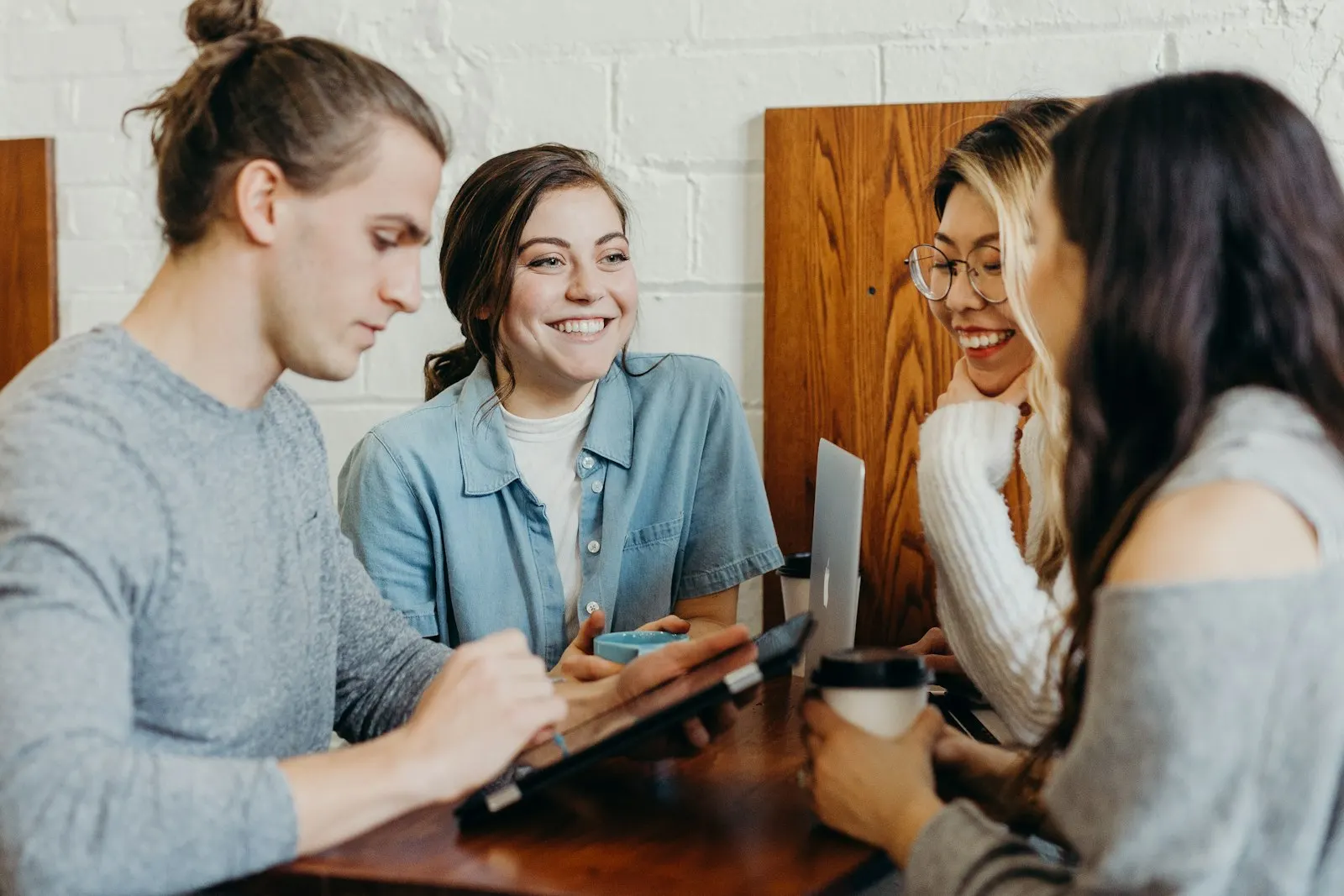 Four people laughing and talking at a wooden cafe table with coffee and a laptop.
