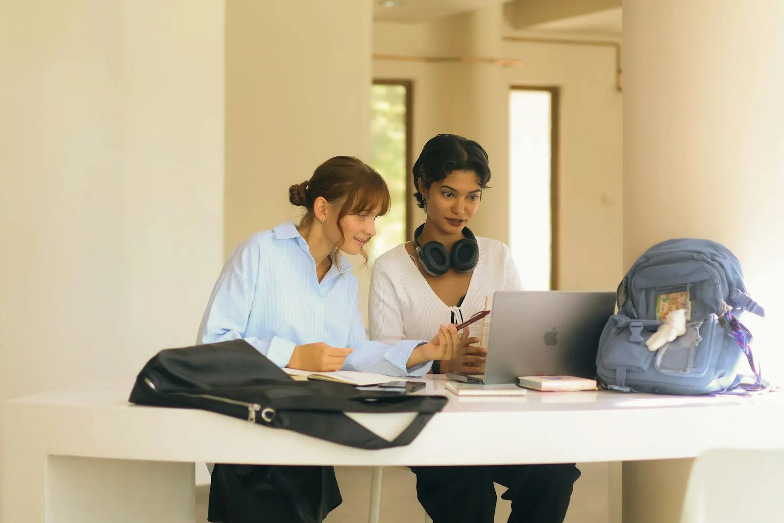 Two students sitting side by side at a table, looking at a laptop and talking.