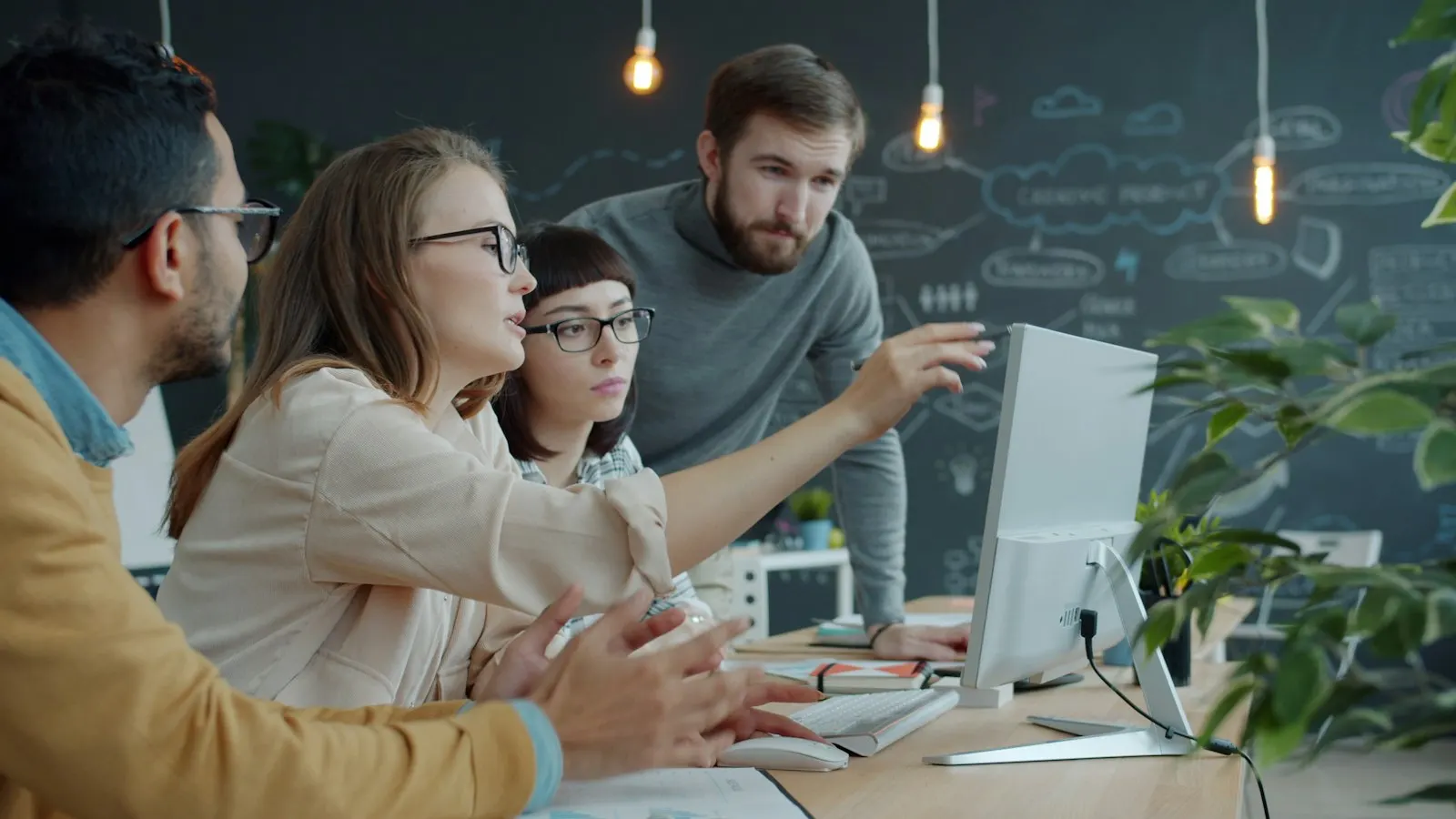 Four colleagues gathered around a computer screen in a bright workspace, one of them pointing at it.