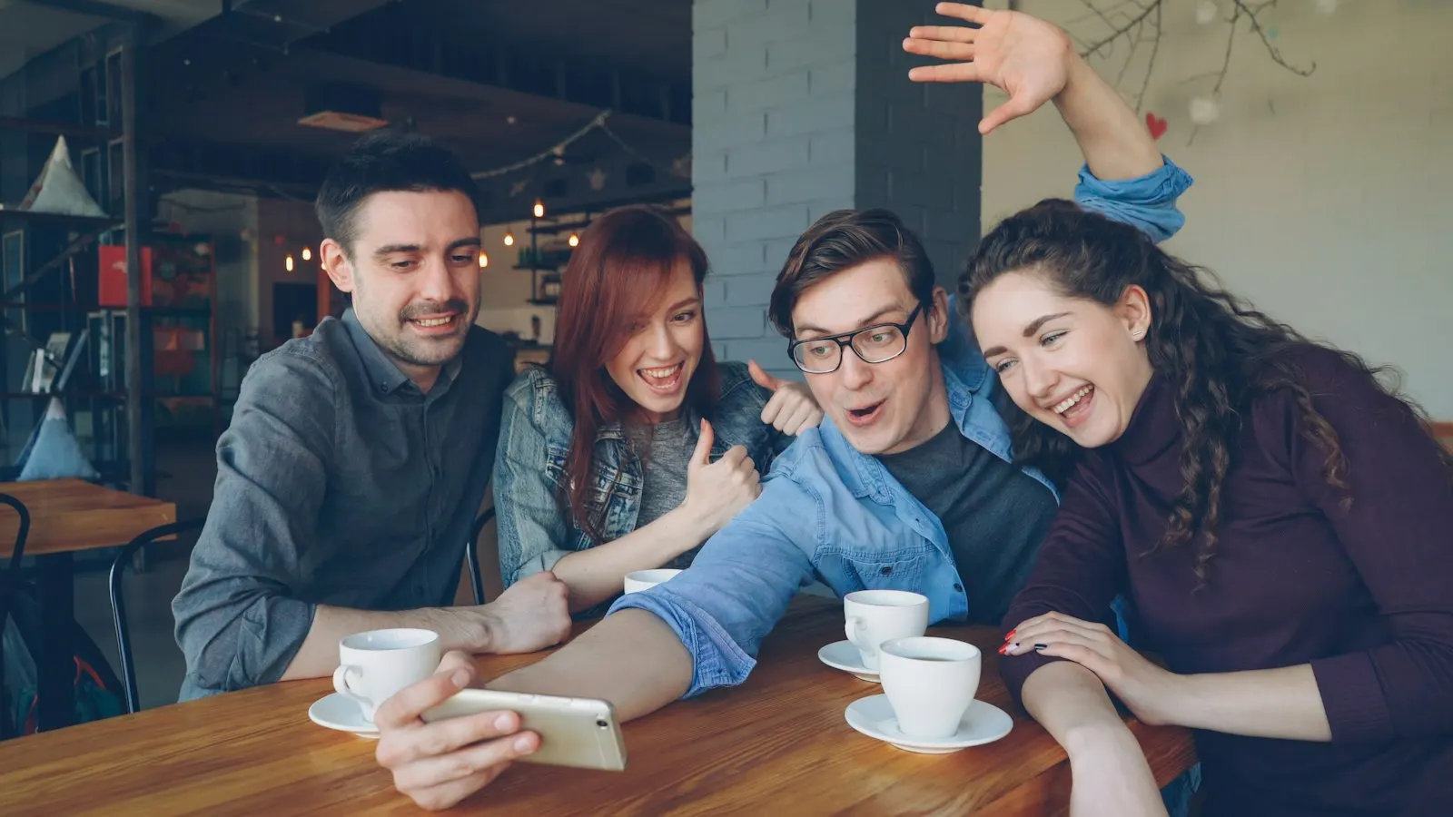 Four friends laughing around a cafe table while one of them takes a photo on a phone.