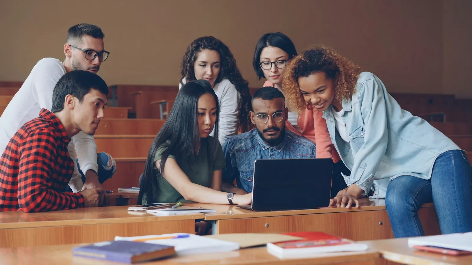 A group of students gathered around a laptop in a lecture hall, looking at the screen together.